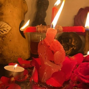 K&ouml;nnte beinhalten: Eine rosa Kerze in Form von zwei sich umarmenden Figuren, platziert in einer dunkelroten Basis, umgeben von roten Rosenbl&auml;ttern. Eine horizontale rote Kerze ist dahinter befestigt, mit einer brennenden vertikalen roten Kerze und zwei Teelichtern.