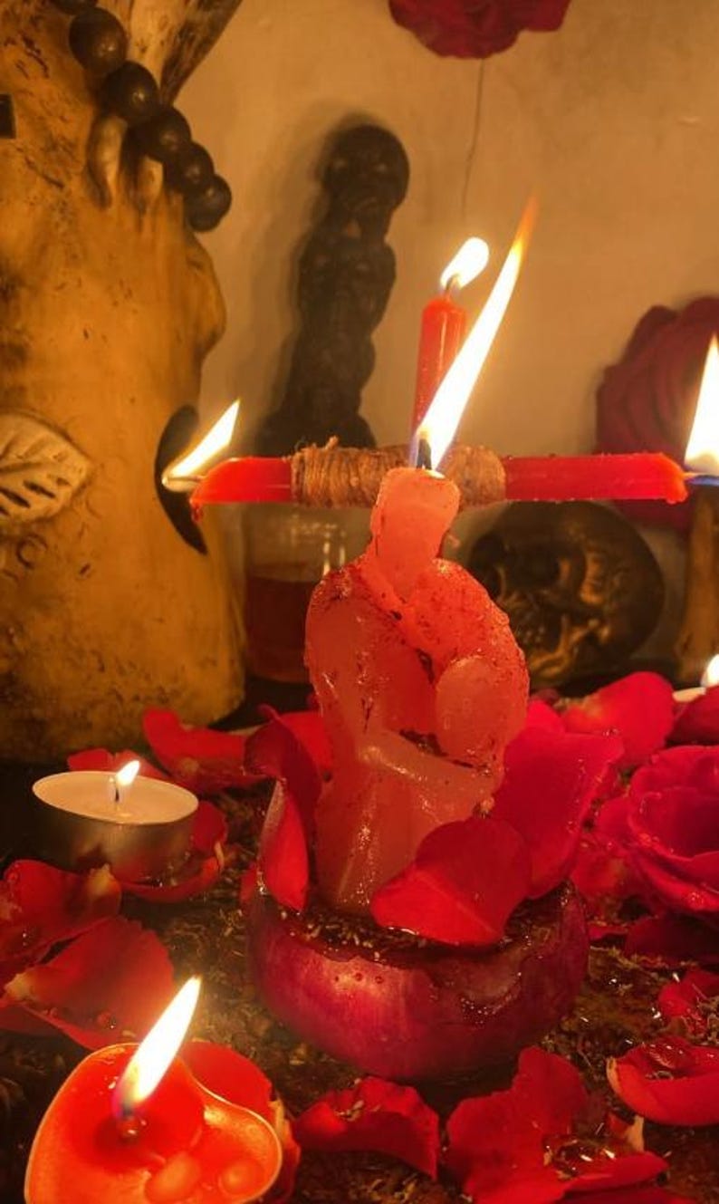 K&ouml;nnte beinhalten: Eine rosa Kerze in Form von zwei sich umarmenden Figuren, platziert in einer dunkelroten Basis, umgeben von roten Rosenbl&auml;ttern. Eine horizontale rote Kerze ist dahinter befestigt, mit einer brennenden vertikalen roten Kerze und zwei Teelichtern.