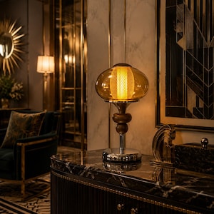 Puede incluir: Una lámpara de mesa con una pantalla de cristal ámbar redondeada, una fuente de luz central de malla cilíndrica y un tallo de madera oscura con detalles esféricos, sobre una base de cromo pulido que refleja la superficie de mármol oscuro.