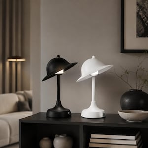 Puede incluir: Dos lámparas de mesa, una en negro mate y otra en blanco mate, cada una con un vástago vertical delgado, una pantalla ancha en forma de cúpula inclinada hacia abajo y una pequeña fuente de luz iluminada situada bajo el centro de la pantalla.