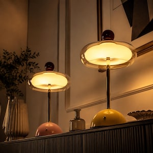 May include: Two table lamps with gold-toned metal stems and dome-shaped bases, one in pastel pink and one in yellow. Each lamp features a clear glass outer shade surrounding a glowing white inner light, topped with a dark brown spherical finial.