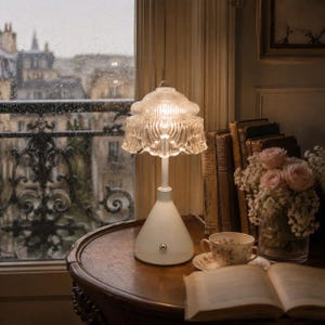 May include: A table lamp with a white conical base and a clear, ribbed glass shade. The lamp features a small metallic power button on the front of the base. The glass shade has a scalloped edge and a textured, fluted design that diffuses light.