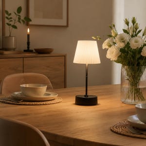 May include: A portable table lamp with a black cylindrical base and a thin black stem supporting a white, pleated, conical lampshade. The lamp emits a warm, soft glow, illuminating the surrounding wooden table surface in a dimly lit room.