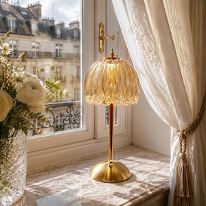 Può includere: Una lampada da tavolo con base in metallo dorato e stelo verticale che sostiene un paralume in vetro color ambra smerlato con una texture a coste. La lampada presenta una base arrotondata in oro lucido e un piccolo pomello decorativo.