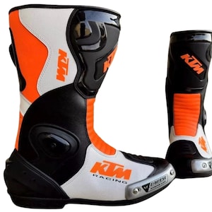 May include: A pair of motorcycle racing boots featuring a colour scheme of black, white, and bright orange. The boots display the KTM logo in orange and the text KTM RACING on the side. A silver-toned protective toe slider is visible on the front.
