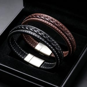 Puede incluir: Dos pulseras de cuero, una negra y otra marrón, con un detalle central trenzado, costuras blancas en contraste a lo largo de los bordes y un cierre magnético rectangular de metal plateado, presentadas dentro de una caja negra.