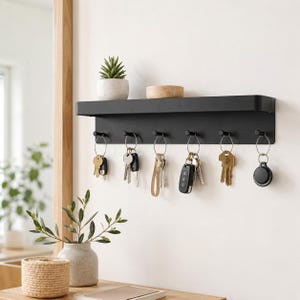 Op de afbeelding: Een matzwarte wandplank met zes geïntegreerde haken voor het ophangen van voorwerpen. Op de plank staan een kleine vetplant in een pot en een ronde houten kom. Zes metalen sleutelringen met diverse sleutels hangen aan de haken.