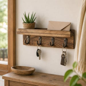May include: A wall-mounted wooden shelf with four dark metal hooks. The shelf holds a small potted succulent and a tan envelope. Two sets of metal keys hang from the hooks, one with a dark leather fob. The wood has a visible grain and brown finish.