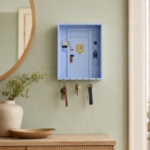 Puede incluir: Un llavero de pared azul claro con forma de puerta, que presenta un detalle de marco de fotos amarillo, un pequeño icono de bolso azul y un acento a rayas. La unidad rectangular incluye cinco ganchos de metal con tres juegos de llaves.