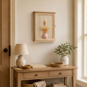 May include: A rectangular light wood wall cabinet with a hinged door featuring a textured beige panel. Inside, a small pink vase holds a bouquet of dried yellow flowers and a single tan plume, creating a decorative display on the wall.