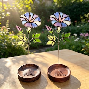 May include: Two stained glass morning glory flowers with pastel blue, pink, and white petals, green leaves, and a pink bud. Each flower is mounted on a thin metal stem attached to a round, dark wood base engraved with the names Anna and Alice.