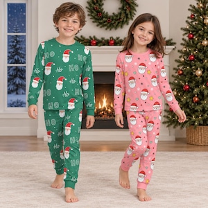 Può includere: Set pigiama a due pezzi con stampa di Babbo Natale e Ho Ho Ho. Un set è verde con testo bianco e volti di Babbo Natale, l'altro è rosa con volti di Babbo Natale, stelle e puntini. Entrambi includono maglie a maniche lunghe e pantaloni abbinati.