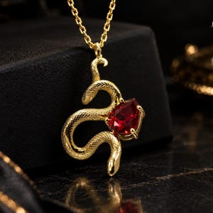 May include: A gold-toned necklace featuring a textured snake pendant coiled around a pear-shaped, deep red gemstone. The pendant hangs from a delicate gold chain, showcasing intricate scale detailing on the snake's body and a prong setting.