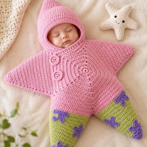 May include: A pink crocheted star-shaped baby bunting with a hood, two round buttons, and light green lower points featuring purple flower-shaped accents. A small, cream-colored crocheted starfish toy with a smiling face is positioned nearby.