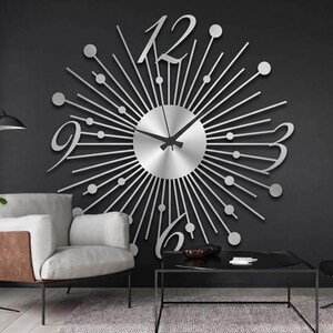 Puede incluir: Un gran reloj de pared plateado con dise&ntilde;o de rayos de sol, que presenta un dial central circular, radios met&aacute;licos finos rematados con peque&ntilde;as esferas y grandes n&uacute;meros estilizados en las posiciones de las doce, tres, seis y nueve, con manecillas negras.