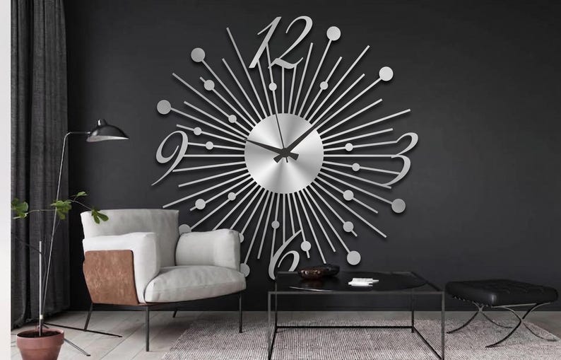 Puede incluir: Un gran reloj de pared plateado con dise&ntilde;o de rayos de sol, que presenta un dial central circular, radios met&aacute;licos finos rematados con peque&ntilde;as esferas y grandes n&uacute;meros estilizados en las posiciones de las doce, tres, seis y nueve, con manecillas negras.