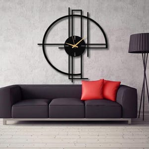 May include: A large black wall clock featuring a circular frame intersected by vertical and horizontal lines. The clock face is a solid black circle with gold-coloured hour and minute hands, mounted on a wall above a dark sofa with red cushions.
