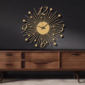 Puede incluir: Un reloj de pared dorado con dise&ntilde;o de rayos de sol, que presenta una esfera circular central con radios finos, peque&ntilde;os acentos circulares en las puntas y n&uacute;meros grandes estilizados para el 12, 3, 6 y 9. Tiene dos manecillas negras en el centro.