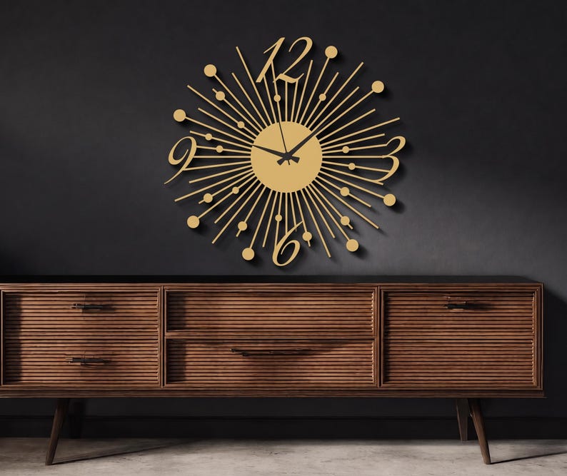 Puede incluir: Un reloj de pared dorado con dise&ntilde;o de rayos de sol, que presenta una esfera circular central con radios finos, peque&ntilde;os acentos circulares en las puntas y n&uacute;meros grandes estilizados para el 12, 3, 6 y 9. Tiene dos manecillas negras en el centro.