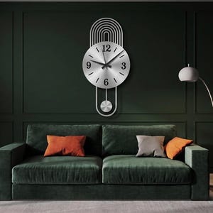 May include: A silver-toned metal wall clock featuring a circular face with black numerals at the three, six, nine, and twelve o'clock positions. The clock has a vertical design with arched lines above the face and a pendulum hanging below.