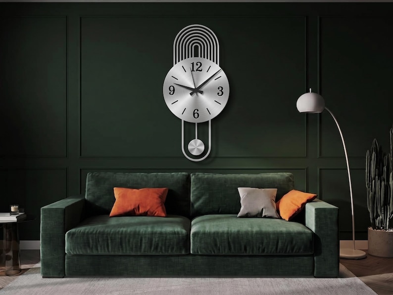 May include: A silver-toned metal wall clock featuring a circular face with black numerals at the three, six, nine, and twelve o'clock positions. The clock has a vertical design with arched lines above the face and a pendulum hanging below.