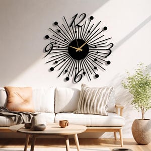 Puede incluir: Un reloj de pared negro con dise&ntilde;o de rayos de sol, centro circular, radios met&aacute;licos finos rematados con peque&ntilde;as esferas y grandes n&uacute;meros estilizados para el 3, 6, 9 y 12. Las manecillas doradas contrastan con el marco de color negro.