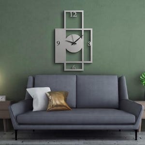 Pu&ograve; includere: Un orologio da parete rettangolare in metallo argentato con una cornice geometrica a sezioni aperte e un quadrante circolare centrale. Il quadrante mostra i numeri 3, 6, 9 e 12 in un semplice carattere sans-serif con due sottili lancette nere.