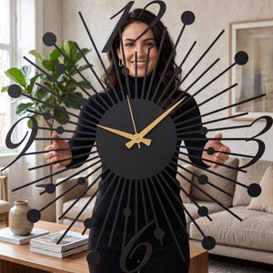 Puede incluir: Un gran reloj de pared negro con dise&ntilde;o de rayos de sol, centro circular y manecillas doradas. La esfera muestra n&uacute;meros negros estilizados en las posiciones de las doce, tres, seis y nueve, rodeados por radios met&aacute;licos que terminan en peque&ntilde;as esferas negras.