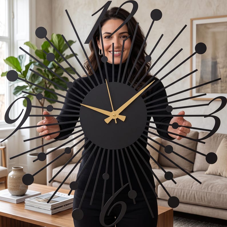 Puede incluir: Un gran reloj de pared negro con dise&ntilde;o de rayos de sol, centro circular y manecillas doradas. La esfera muestra n&uacute;meros negros estilizados en las posiciones de las doce, tres, seis y nueve, rodeados por radios met&aacute;licos que terminan en peque&ntilde;as esferas negras.