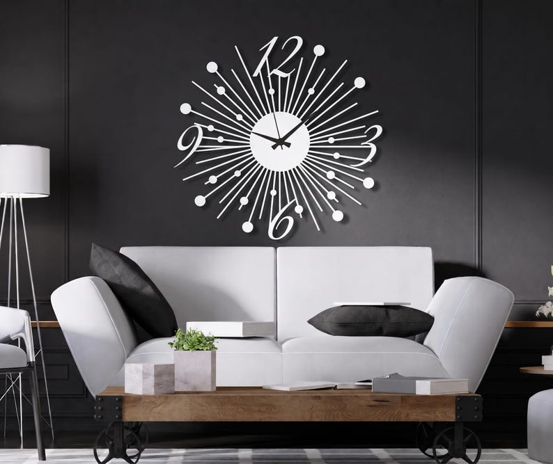 Puede incluir: Un reloj de pared blanco con dise&ntilde;o de rayos de sol, que presenta una esfera central circular con manecillas negras, rodeada de finos radios met&aacute;licos rematados con peque&ntilde;os c&iacute;rculos y grandes n&uacute;meros estilizados en las posiciones de las doce, tres, seis y nueve.