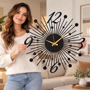 Puede incluir: Un reloj de pared negro con dise&ntilde;o de rayos de sol, centro circular, radios met&aacute;licos finos rematados con peque&ntilde;as esferas y grandes n&uacute;meros estilizados para el 12, 3, 6 y 9. El reloj tiene tres manecillas finas de color dorado.