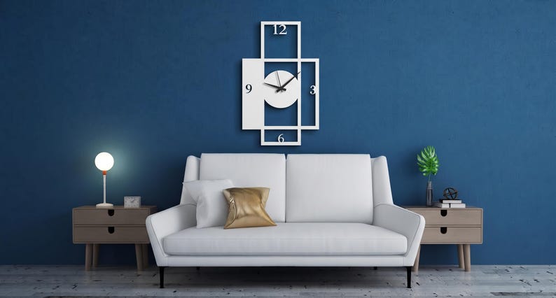 Pu&ograve; includere: Un orologio da parete bianco con una cornice geometrica composta da contorni rettangolari e un quadrante centrale circolare. Il quadrante mostra i numeri 3, 6, 9 e 12 in un carattere nero senza grazie, con sottili lancette nere.