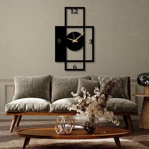 Pu&ograve; includere: Un orologio da parete nero con un design geometrico di cornici rettangolari sovrapposte. Il quadrante &egrave; un cerchio nero pieno con lancette dorate per ore e minuti. I numeri 12, 3, 6 e 9 sono posizionati all'interno della struttura.