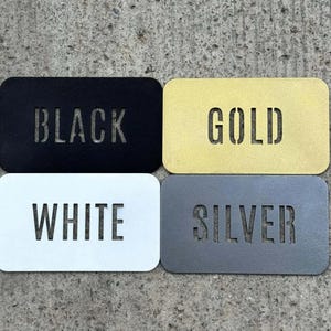 Puede incluir: Cuatro placas met&aacute;licas rectangulares con esquinas redondeadas, cada una con una palabra cortada con l&aacute;ser. La placa superior izquierda es negra con la palabra BLACK, la superior derecha dorada con GOLD, la inferior izquierda blanca con WHITE y la inferior derecha plateada con SILVER.