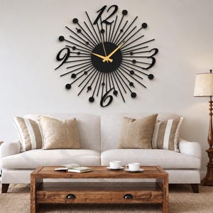 Puede incluir: Un gran reloj de pared negro con dise&ntilde;o de rayos de sol, centro circular, radios finos rematados con peque&ntilde;os c&iacute;rculos negros y cuatro grandes n&uacute;meros estilizados en las posiciones de las doce, tres, seis y nueve. Tiene manecillas doradas.