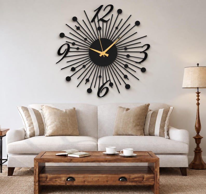 Puede incluir: Un gran reloj de pared negro con dise&ntilde;o de rayos de sol, centro circular, radios finos rematados con peque&ntilde;os c&iacute;rculos negros y cuatro grandes n&uacute;meros estilizados en las posiciones de las doce, tres, seis y nueve. Tiene manecillas doradas.