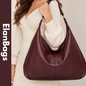 Peut inclure: Un grand sac hobo bordeaux profond avec une surface texturée, une bandoulière unique et une couture verticale centrale. Le sac présente des détails métalliques argentés reliant la sangle au corps. Le texte ElanBags est visible à gauche.