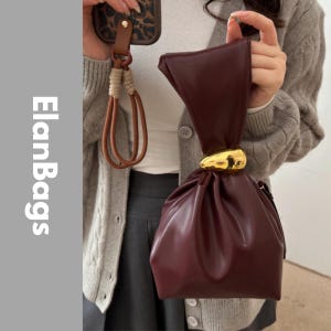May include: A dark burgundy handbag with a smooth finish, featuring a gathered top design secured by a polished gold-tone metallic accent. The bag has a soft, slouchy structure and includes the text ElanBags vertically on the left side.
