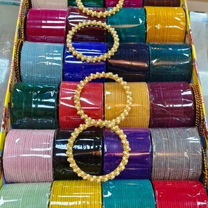 Op de afbeelding: Een rechthoekige doos met meerdere sets armbanden in diverse effen kleuren, waaronder rood, blauw, groen, geel, paars, roze en grijs. Drie dunne goudkleurige armbanden versierd met kleine witte kraaltjes liggen bovenop de stapels.
