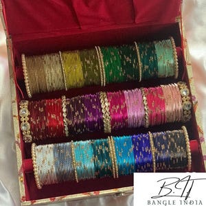 Op de afbeelding: Een verzameling veelkleurige metalen armbanden gerangschikt in drie rijen in een doos met rode voering. De armbanden hebben metallic afwerkingen in goud, groen, rood, paars, roze, blauw en zilver, sommige geaccentueerd met kleine witte kralen.