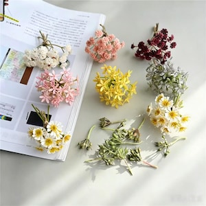 Op de afbeelding: Een verzameling kleine, kunstmatige bloemenboeketten in diverse kleuren, waaronder wit, lichtroze, donkerrood, geel en zilver, samen met verschillende groene, ongeopende bloemknoppen, gerangschikt op een oppervlak naast een open tijdschrift.