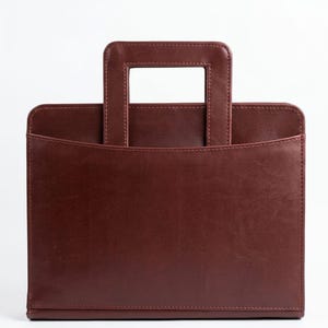 May include: A dark burgundy rectangular briefcase with a smooth, leather-like finish. It features a structured design with a single integrated rectangular handle at the top and a curved front pocket opening that spans the width of the bag.
