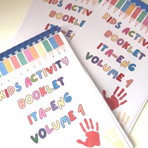 May include: Three spiral-bound activity booklets with white covers featuring a colourful pencil border at the top. The text reads KIDS ACTIVITY BOOKLET ITA-ENG VOLUME 1 in multi-coloured letters, accompanied by red, yellow, and blue handprint graphics.