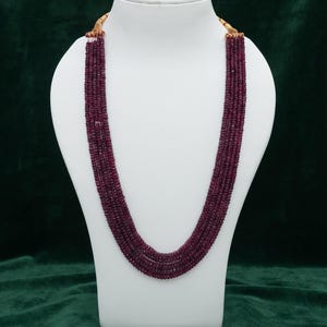 May include: A multi-strand necklace featuring numerous small, faceted, deep red ruby beads arranged in several parallel rows. The strands are gathered at the top with gold-toned metal caps and secured with a light-colored cord closure.
