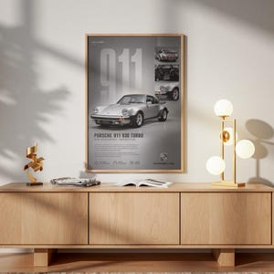 May include: A framed vertical poster featuring a silver Porsche 911 930 Turbo, the text 911, and the Porsche logo. The poster includes smaller inset images of the car, technical specifications, and descriptive text on a grey background.