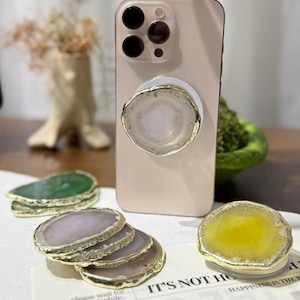 May include: A light pink smartphone with a white circular phone grip featuring a translucent white agate slice with a gold-toned metallic edge. Several additional agate slice phone grips in green, grey, and yellow are arranged on a white surface.