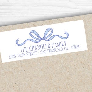 May include: A white rectangular label with a light blue bow illustration and text. The text reads "THE CHANDLER FAMILY" and the address "2528 UNION STREET - SAN FRANCISCO, CA - 94123". The label is on a light brown envelope.