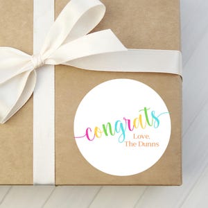 May include: A white round gift tag with the word "congrats" in rainbow colors and the text "Love, The Dunns" in orange. The tag is attached to a brown gift box with a white ribbon.