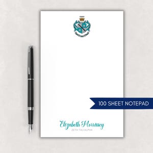 May include: A white notepad with a teal and black crest and the name "Elizabeth Morrissey" in teal script. A black pen is to the left of the notepad. A dark blue banner reads "100 SHEET NOTEPAD".