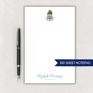 May include: A white notepad with a blue and gold crest at the top and a name in light blue script at the bottom. A black pen is to the left of the notepad. A blue banner reads "100 SHEET NOTEPAD."